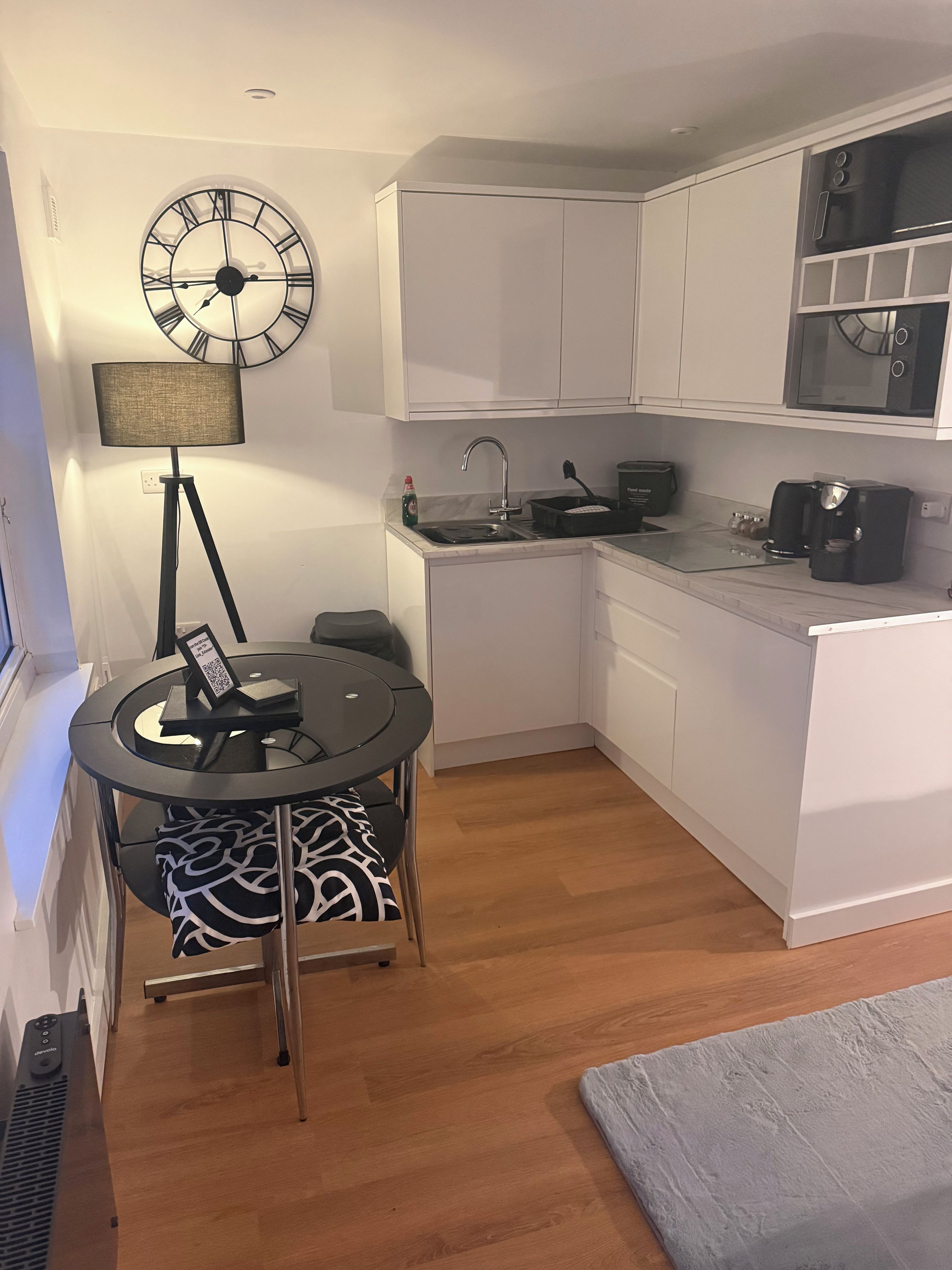A compact kitchen with white cabinets, a round clock, a small dining table with chairs, a standing lamp, and a built-in microwave.