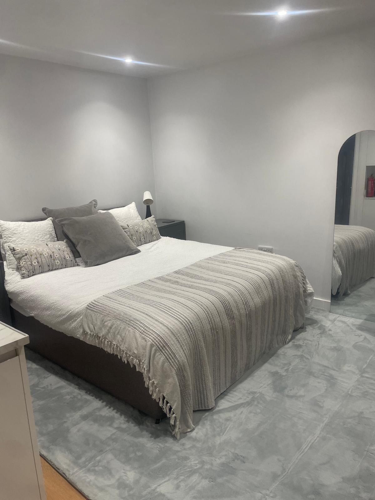 A neatly arranged bedroom with a large bed, a bedside table, a lamp, and a mirror.