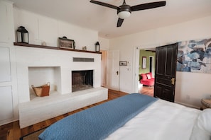 A bedroom with a white brick fireplace, a ceiling fan, a bed with a blue quilt, and a red chair in the background.