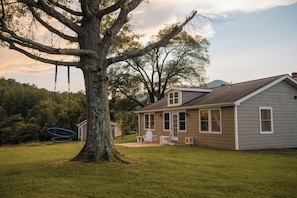 A house with a large tree in the foreground, a swing set, and a patio with outdoor furniture.