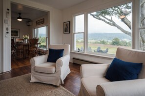 Une pièce comprenant deux fauteuils blancs, une table à manger avec des chaises, et de grandes fenêtres donnant sur l’extérieur.