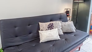 A dark-colored sofa with multiple cushions, a framed landscape painting, and a floor lamp in a room with white walls and tiled flooring. - SHB - Orquidário 3 min Praia em Studio (Santos)