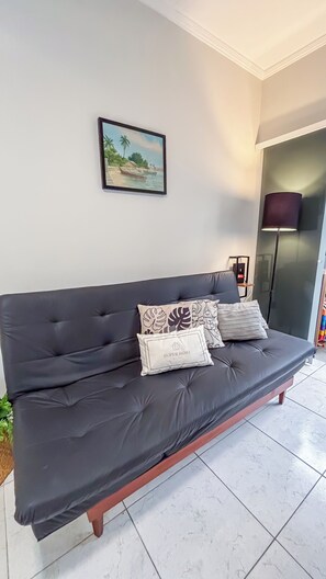 Um sofá escuro com várias almofadas, um quadro emoldurado de uma paisagem e um abajur em um quarto com paredes brancas e piso de azulejos.