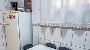A compact kitchen with a white refrigerator, a small dining table with four chairs, and a window with a sheer curtain. - SHB - Orquidário 3 min Praia em Studio (Santos)