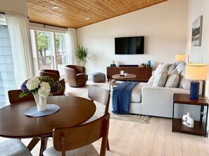 A modern living room with a wooden ceiling, a flat-screen TV mounted on the wall, a sofa, a coffee table, and a dining area with a table and chairs. - Lake Sawyer Waterfront Retreat with Private Dock, Near Skiing and Mt. Rainier (Black Diamond)