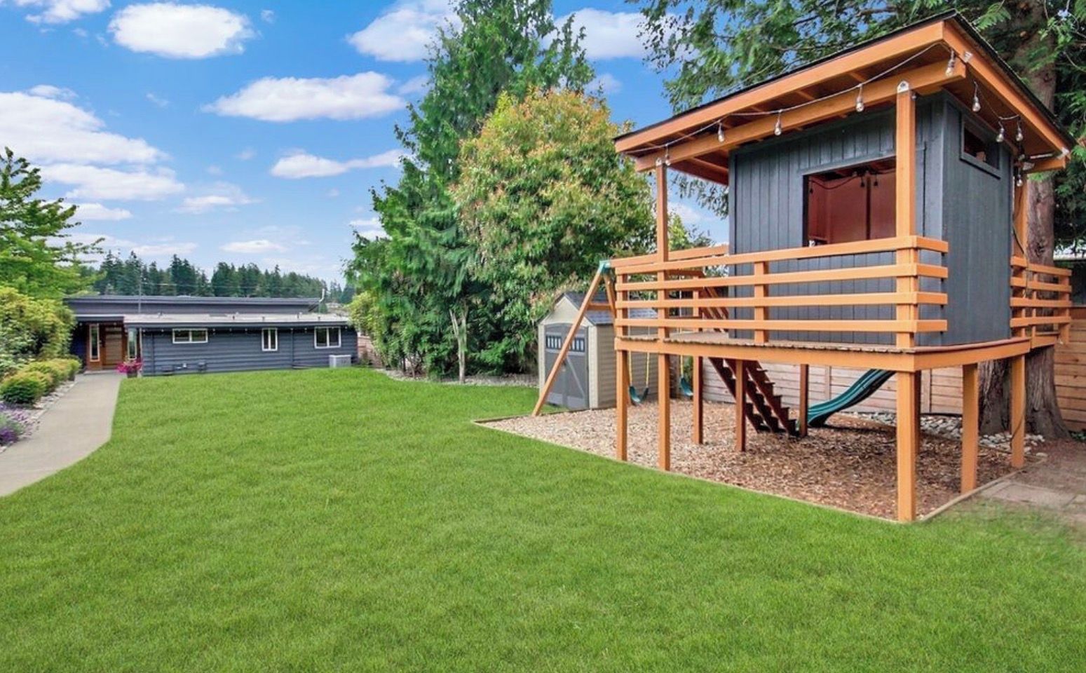 Une propriété résidentielle dotée d’une pelouse bien entretenue, d’une cabane en bois avec un toboggan, et d’une maison visible au loin.
