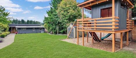 Une propriété résidentielle dotée d’une pelouse bien entretenue, d’une cabane en bois avec un toboggan, et d’une maison visible au loin.