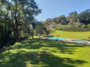 Un césped bien cuidado con zona de piscina, rodeado de árboles y una colina al fondo.