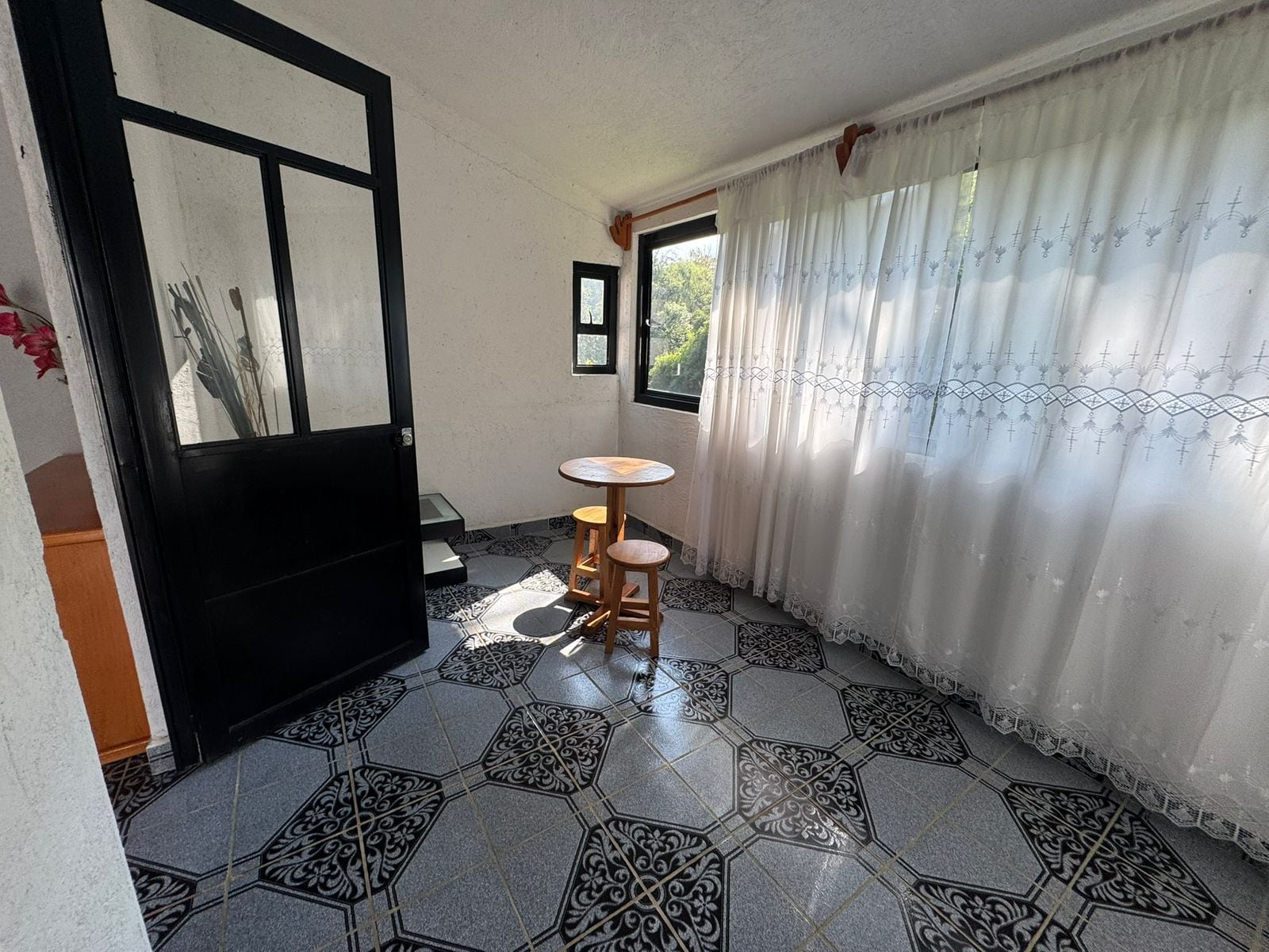 Una habitación con suelo de baldosas, una mesa redonda de madera, una puerta con marco negro y una ventana con vistas a zonas verdes.