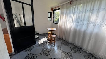 Una habitación con suelo de baldosas, una mesa redonda de madera, una puerta con marco negro y una ventana con vistas a zonas verdes.