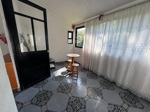 Una habitación con suelo de baldosas, una mesa redonda de madera, una puerta con marco negro y una ventana con vistas a zonas verdes.