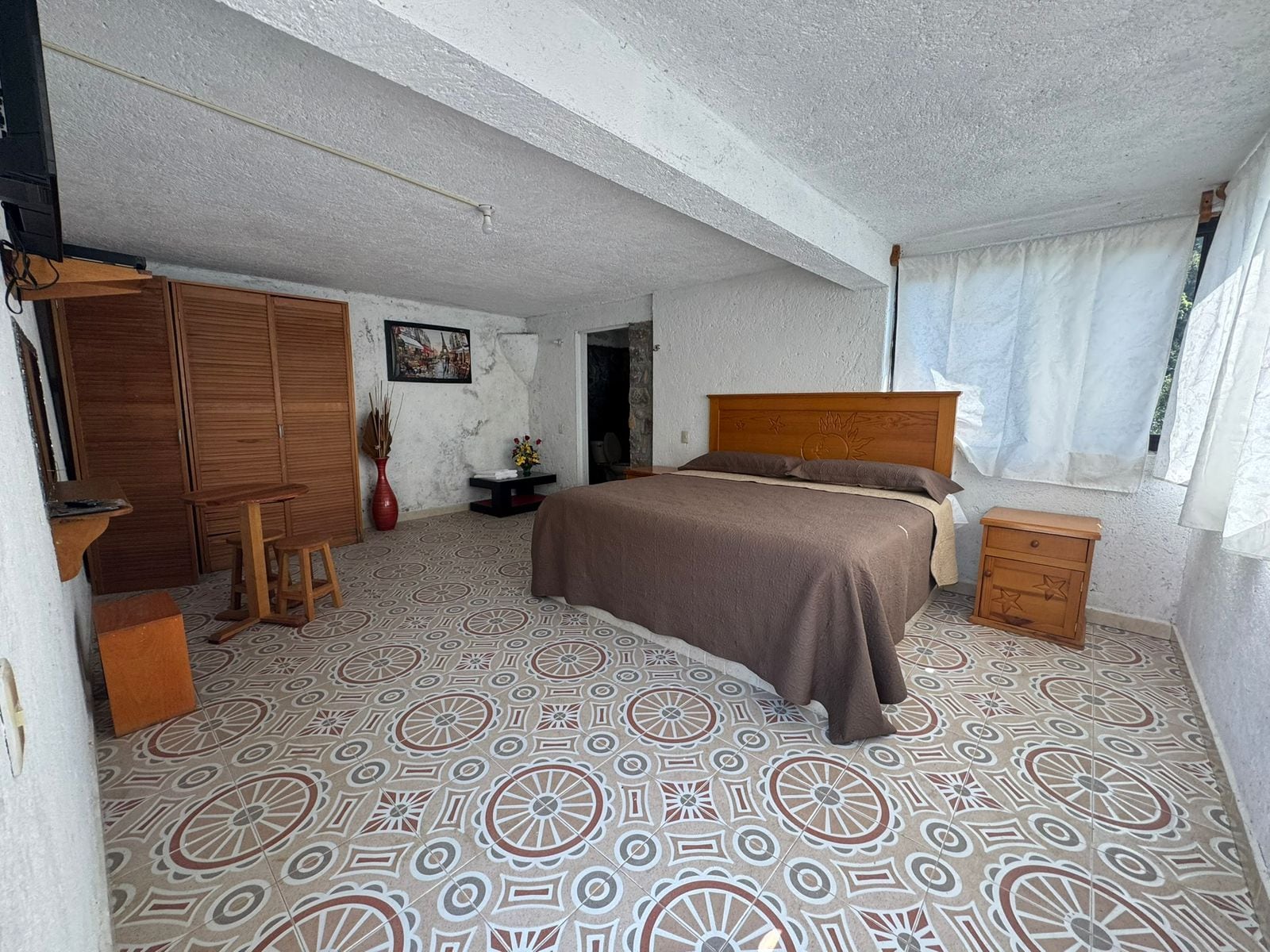 Een slaapkamer met een bed, een houten garderobe, een klein tafeltje en een raam met gordijnen.
