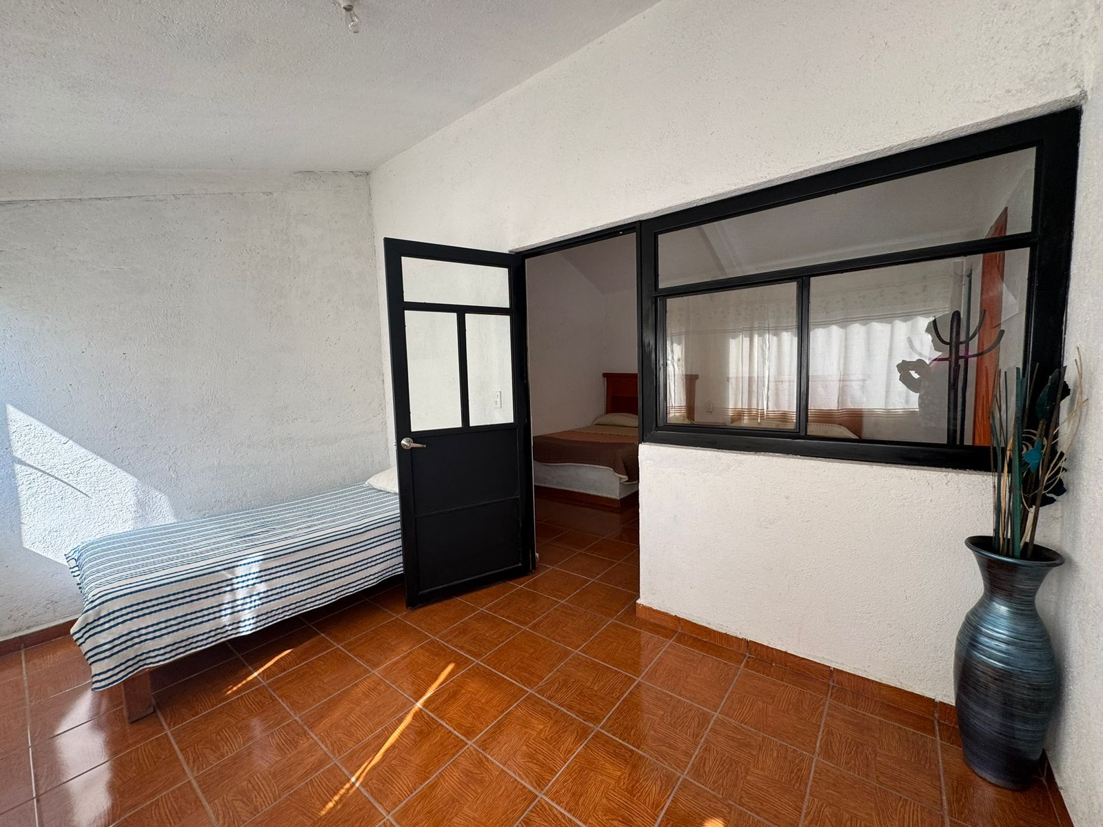 A room with a bed, a black door, a window, and a vase on the floor.