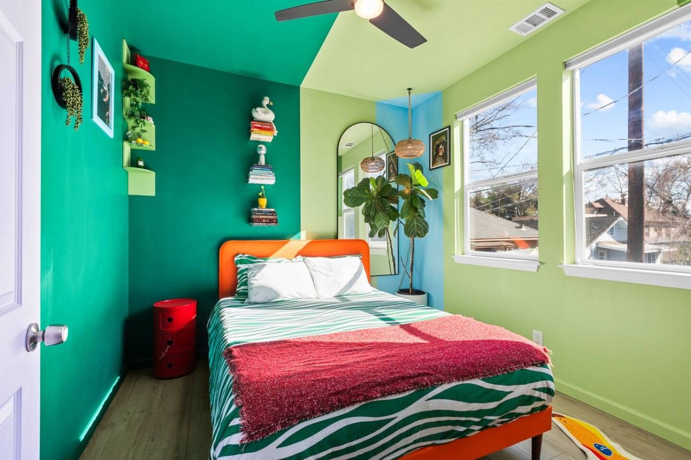 A bedroom with a bed, a red trash bin, a ceiling fan, and a window with a view of the outside.