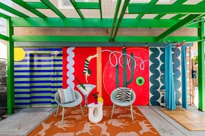 Una zona exterior animada amb una paret mural colorida, dues cadires, una taula petita i un moqueta amb dibuixos.
