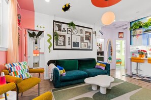 Un saló modern amb un sof à verd, coixins de colors, una taula de café blanca i una decoració vibrant que inclou obra d'art a les parets i plantes.