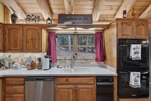 A kitchen with wooden cabinets, a stainless steel dishwasher, a refrigerator, and a window with a view of trees. - Peaceful Log Cabin Getaway on 6 Private Acres (Grass Valley)