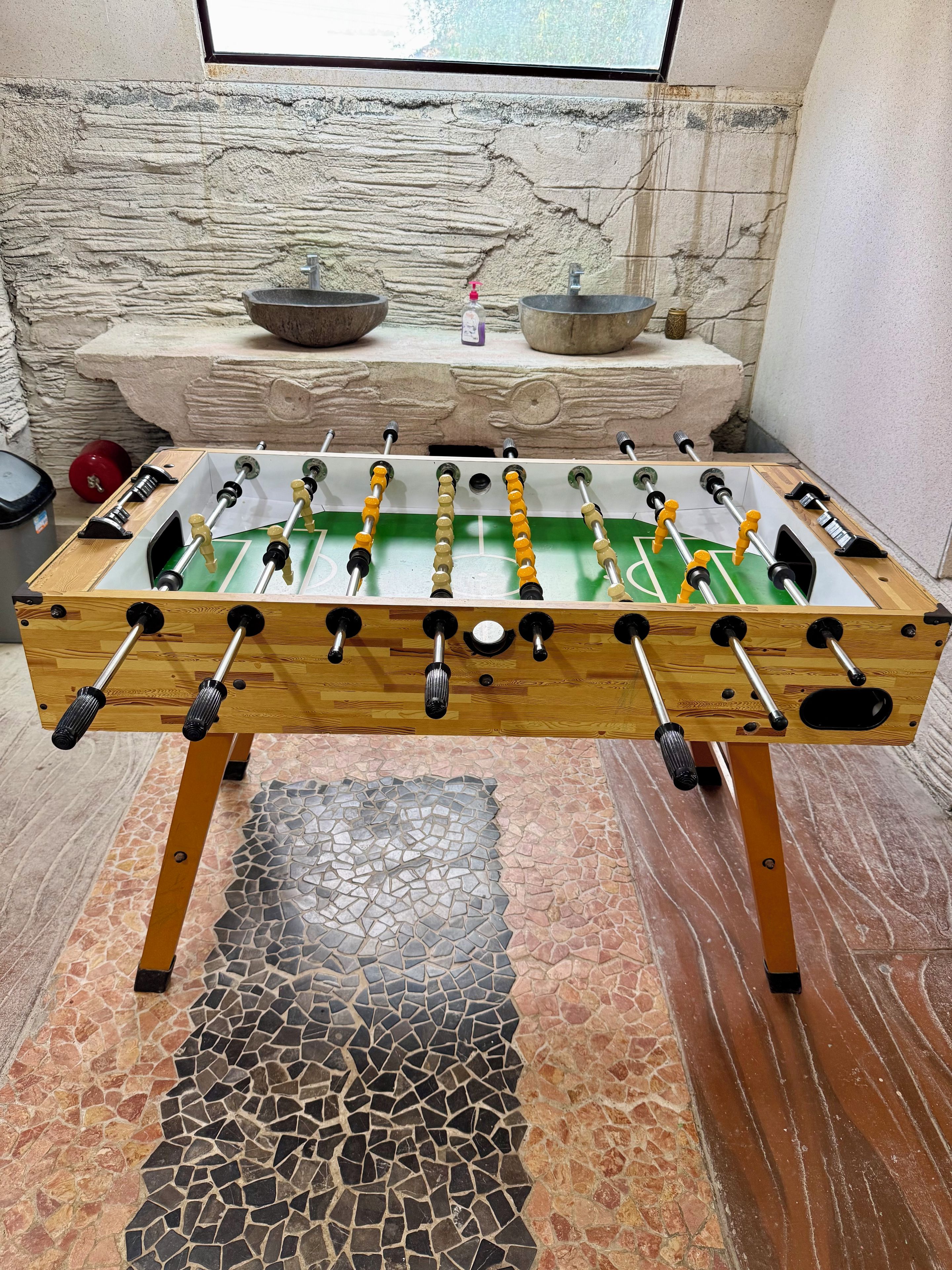 Una mesa de fútbolín con estructura de madera y superficie de juego verde, frente a una pared de piedra con una ventana arriba.