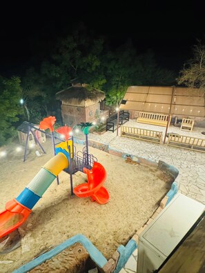A playground with a slide and climbing structures, surrounded by a sandy play area and a building with a thatched roof.
