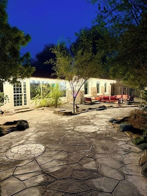 Une terrasse en pierre avec un motif circulaire, un espace détente avec des coussins rouges et un bâtiment doté d’une porte et de fenêtres en verre.