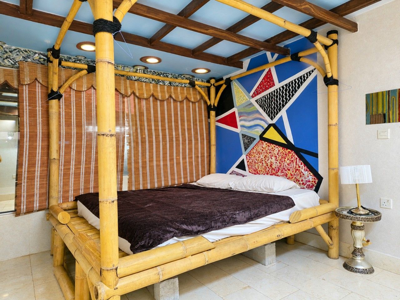 A room with a bed under a bamboo canopy, a patterned wall, and a small table with a lamp.