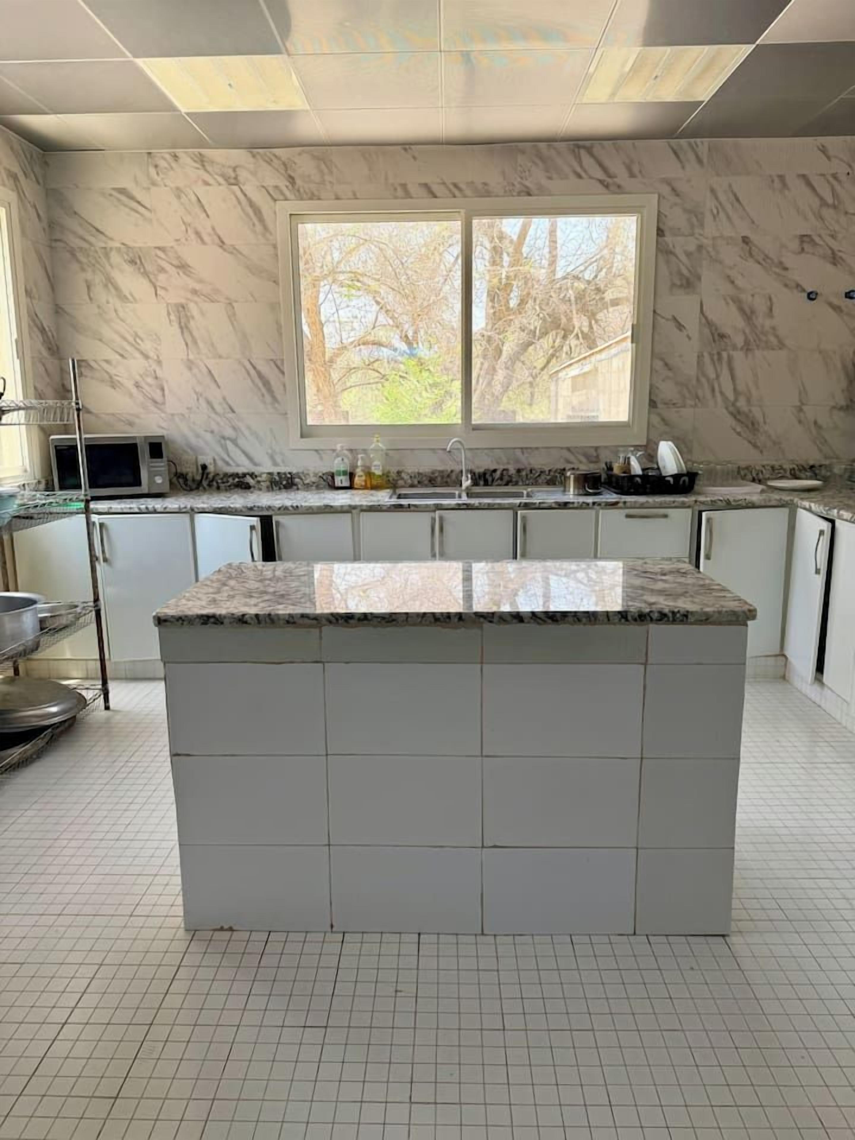 A kitchen with a large island, white tiled floor, and a window with a view of trees.