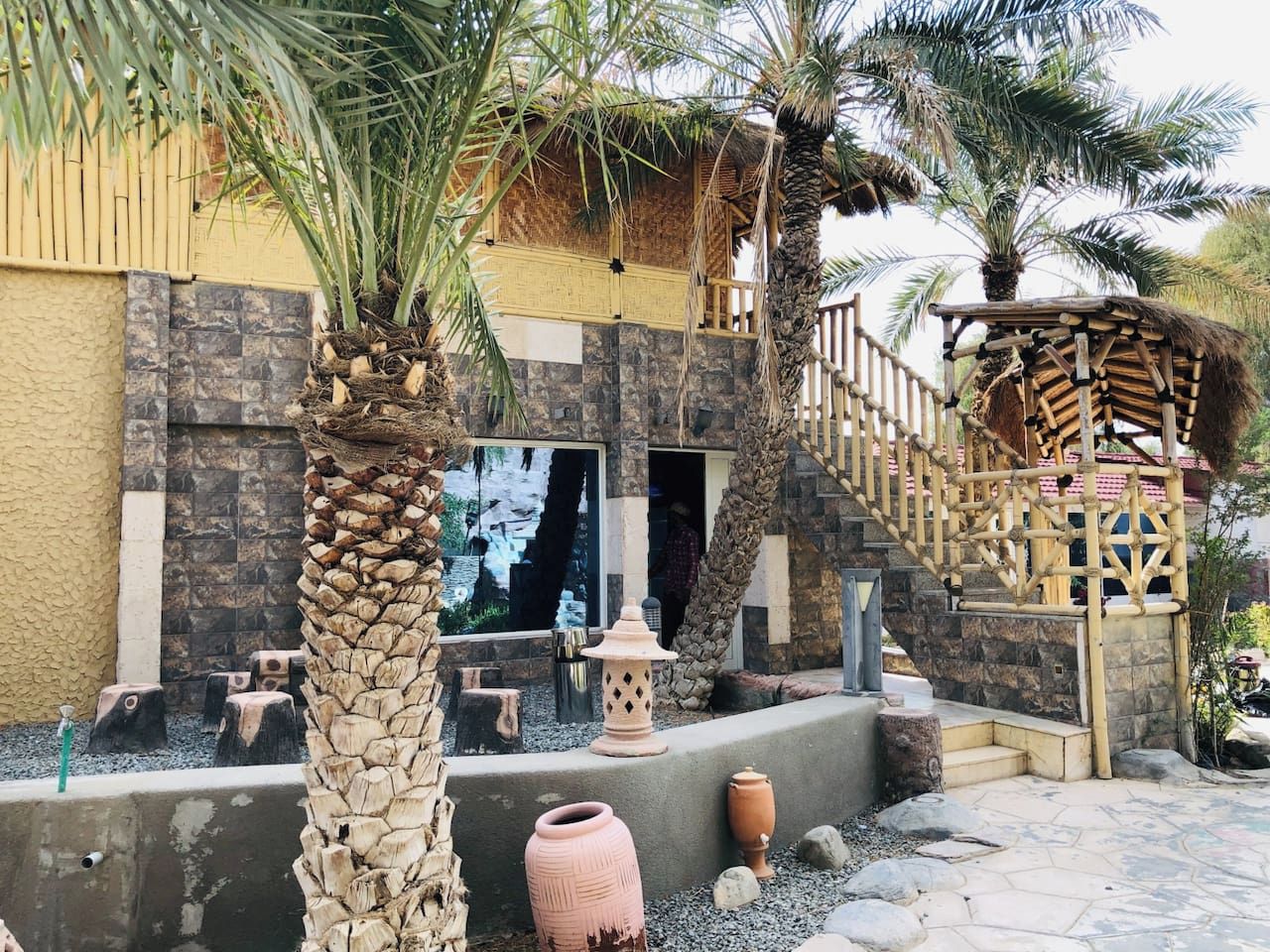 A courtyard with a stone wall, palm trees, a wooden staircase, and a thatched roof structure.
