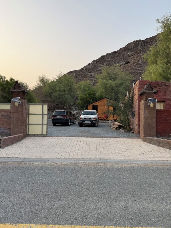 A gated driveway leading to a building with a brick facade, two parked cars, and a mountainous backdrop.
