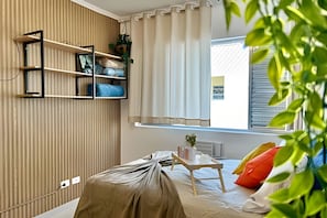 A bedroom with a bed covered in a beige blanket, a bedside table with a small plant, and a window with curtains. - SHB - Studio no Gonzaga à Beira Mar (Santos)