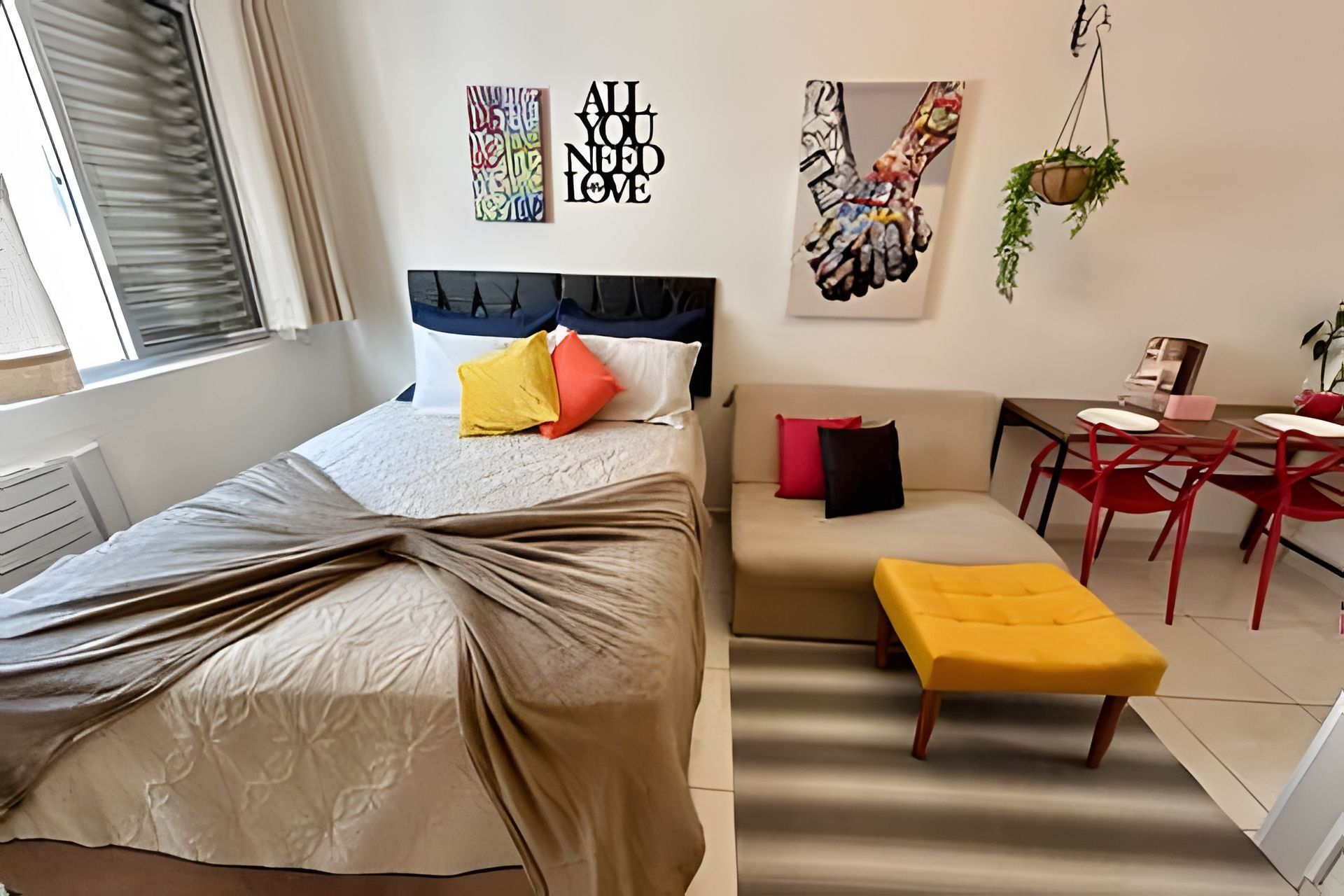 A modern bedroom with a bed, a sofa, and a small table. There are wall art pieces and a plant.