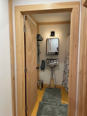 Kapea käytävä johtaa kylpyhuoneeseen, jossa on pesuallas, peili ja pieni hylly, jossa on hygieniatuotteita.