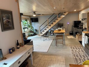 A modern kitchen with a wooden staircase, a dining area with a table and chairs, and a living space with a TV and plants.