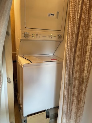 A compact, white washing machine with a control panel, located in a narrow space with a patterned curtain partially visible.