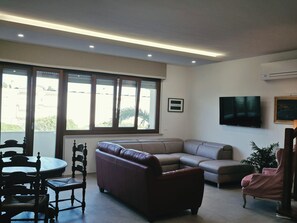 A living room with a brown leather sofa, a grey sofa, a wooden dining table with chairs, a television mounted on the wall, and a large window with blinds. - Zefiro Apartment in Alghero. The perfect place to visit the Coral Riviera (Fertilia)
