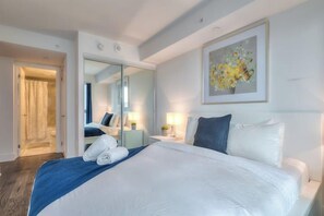 Une chambre d’hôtel avec un grand lit, un miroir, un tableau et une salle de bain visible à travers une porte ouverte.