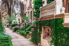 A tree-lined residential street with stone buildings, a wrought iron fence, and a stone lion statue. - Sabal House (Savannah)