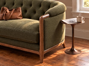 A green upholstered sofa with wooden armrests, a brown leather bag on it, and a small round table with a newspaper and a cup of coffee. - Sabal House (Savannah)