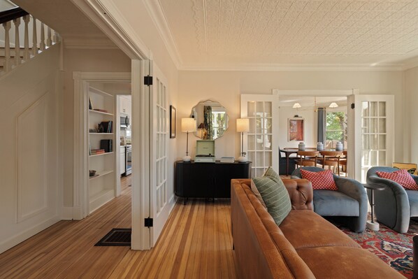 A spacious living room with a brown sofa, a blue armchair, and a patterned rug. There is a dining area with a table and chairs, and a kitchen visible in the background.