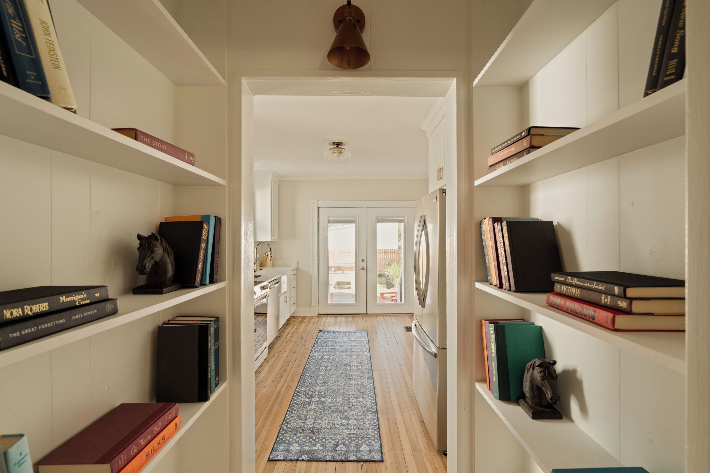 A hallway with bookshelves on both sides, a rug on the floor, and a door at the end leading outside.