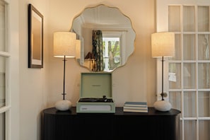 Una habitación con un tocadiscos antiguo sobre una consola oscura, flanqueada por dos lámparas con pantallas texturizadas y un espejo grande arriba.