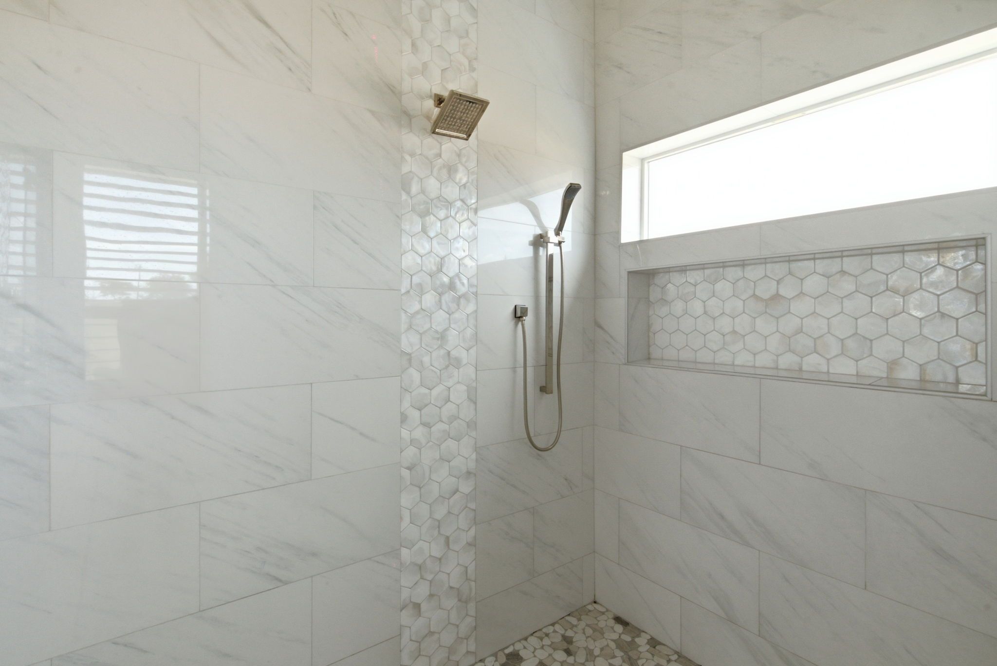 Un baño con baldosas de mármol blanco, una ducha con cabezal de ducha manual y una pared de acento con baldosas hexagonales.