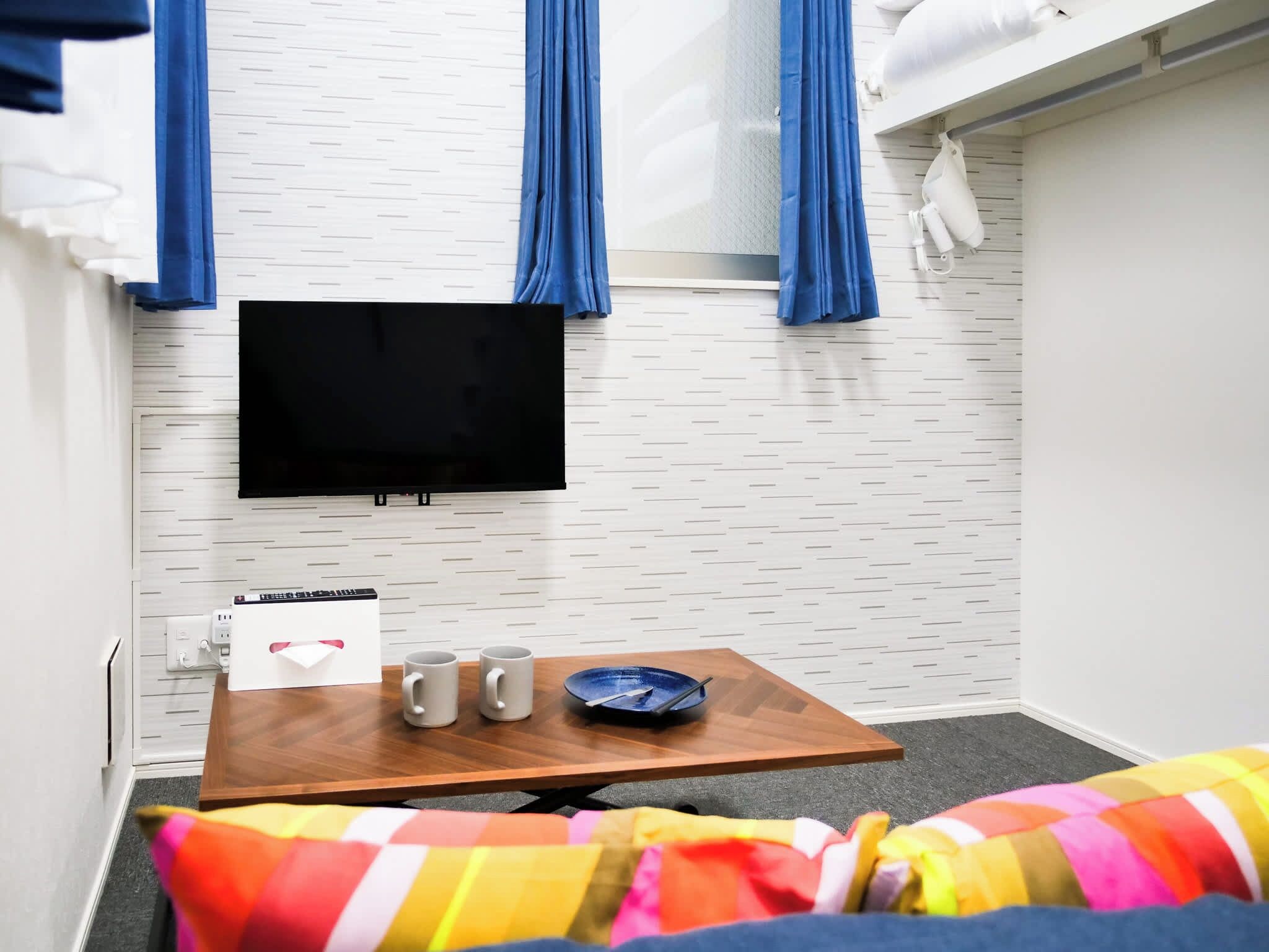 A room with a flat-screen TV, a wooden table with two mugs, a plate, and a colorful striped blanket.