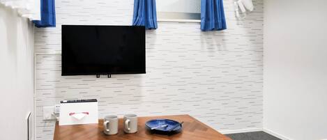 A room with a flat-screen TV, a wooden table with two mugs, a plate, and a colorful striped blanket.