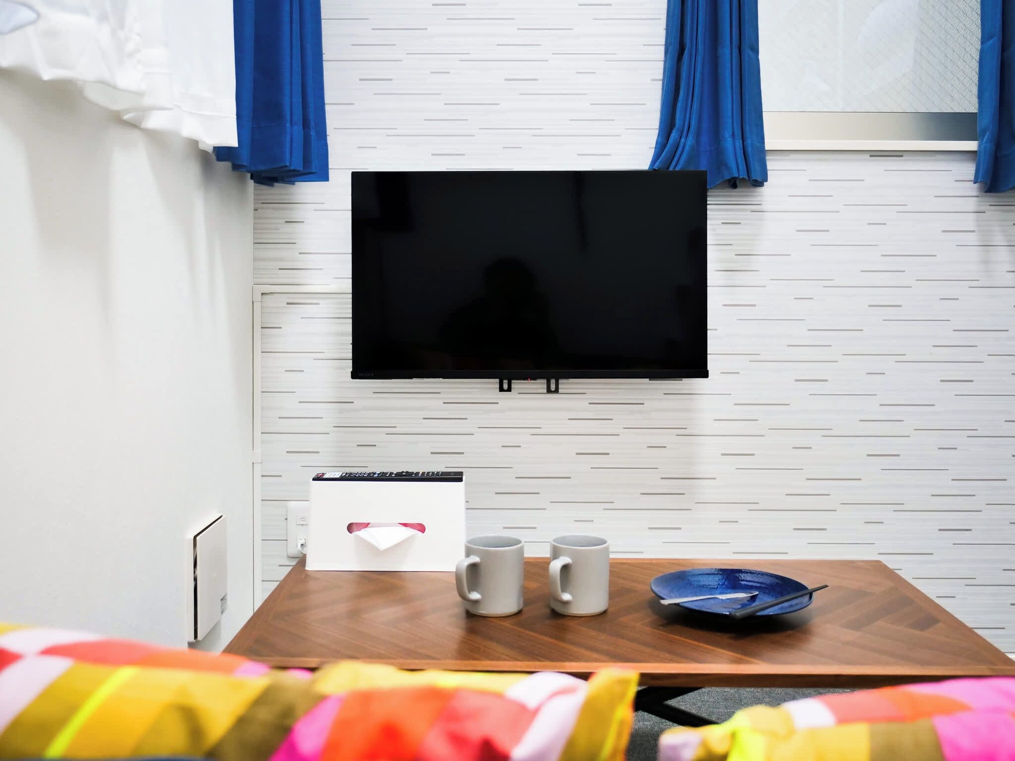 A room with a mounted TV, a small table with three mugs, a blue bowl, and a white box with a red ribbon.