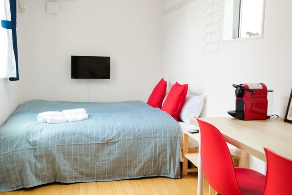 A bedroom with a bed, a TV mounted on the wall, a desk with a chair, and a red suitcase.