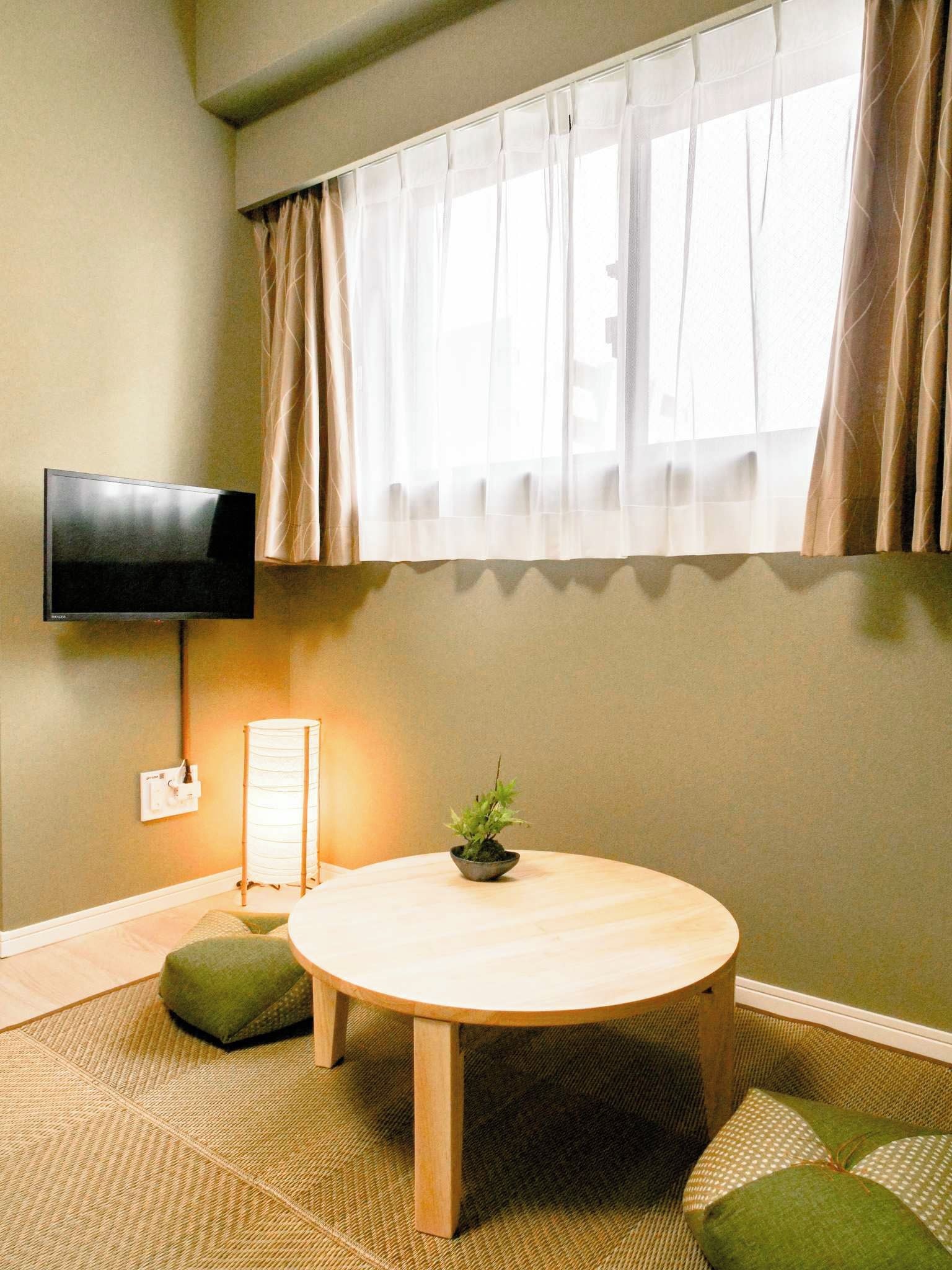A room with a round wooden table, a green cushion, a small potted plant, a flat-screen TV mounted on the wall, and a window with sheer curtains.