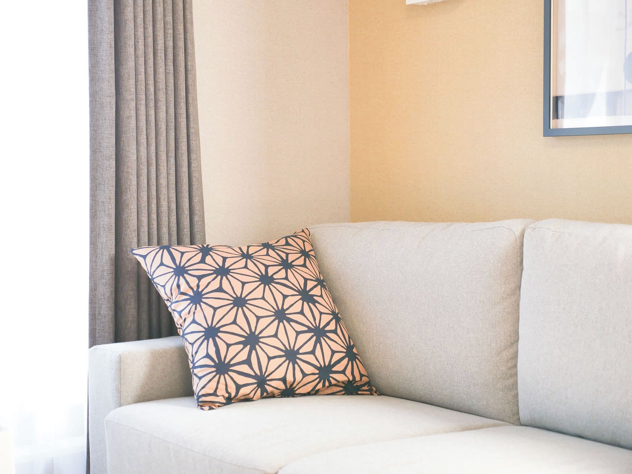 Um canto de quarto com um sofá bege, uma almofada estampada e uma janela com cortinas.