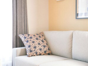 Eine Raumecke mit einem beigen Sofa, einem gemusterten Kissen und einem Fenster mit Vorhängen.