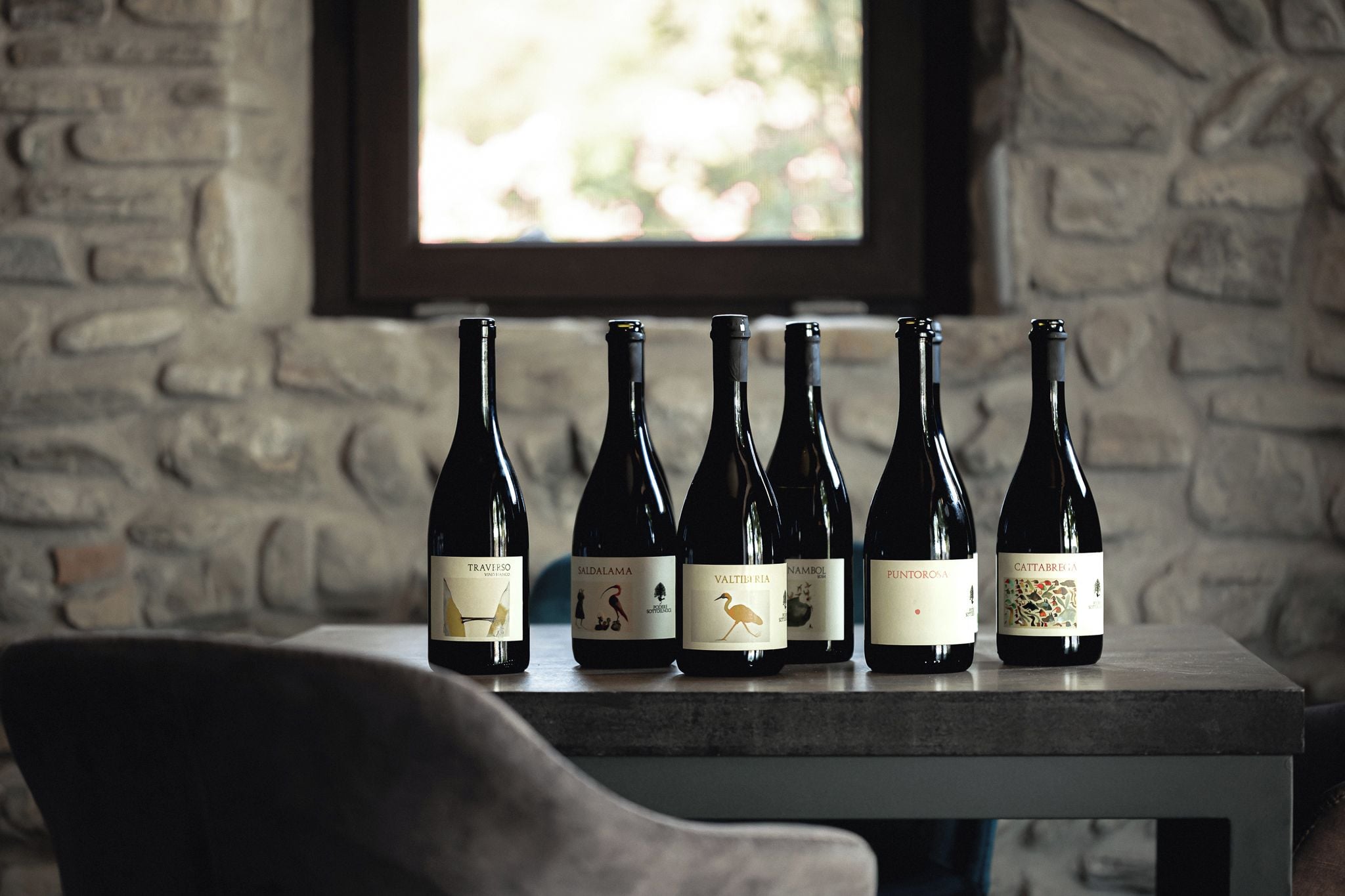 A row of wine bottles on a table with a stone wall and a window in the background.