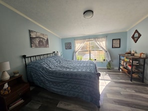 A bedroom with a bed, a nightstand, a dresser, a window with curtains, a bookshelf, and a ceiling light.